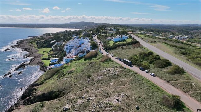 Casapueblo aerial forward push along the Punta Ballena coastline, Uruguay, revealing rocky shoreline, green hillsides, and a road with parked buses and cars.