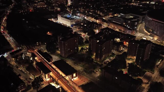 Tottenham residential district glows at night around apartment blocks, illuminated streets and a railway corridor, with dense urban development, drone descending.