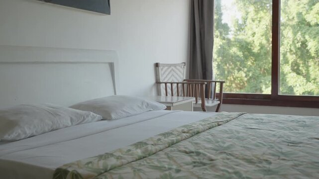 A made bed in a white bedroom with a large window facing green trees