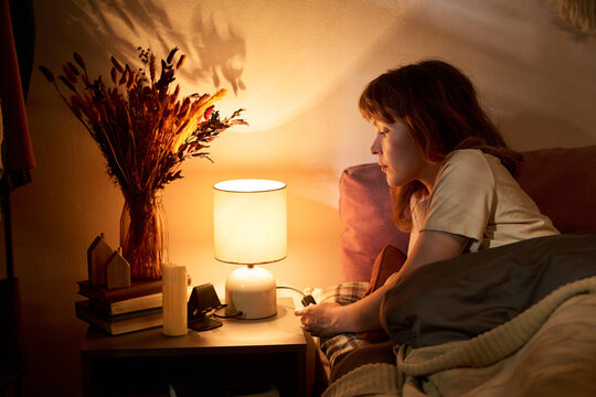 Caucasian young adult woman lying in bed and turning off bedside lamp before sleeping. Image represents healthy sleep hygiene and bedtime routine, ideal for mental wellbeing campaigns