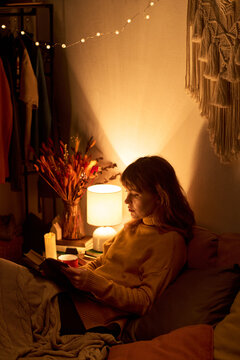 Vertical image of Caucasian young adult woman reading book in bed under warm lamp light, promoting mental wellbeing, self care, and cozy evening relaxation concepts