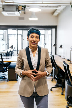 Confident businesswoman with blue hair standing in a modern coworking office. She is collaborating in a startup environment, representing success and professionalism
