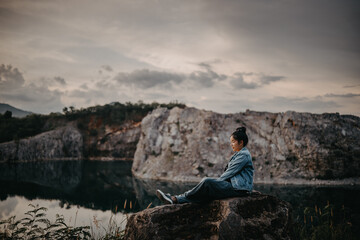 Fototapeta premium Young female tourist sitting on large rock enjoying scenic mountain lake view at dusk.
