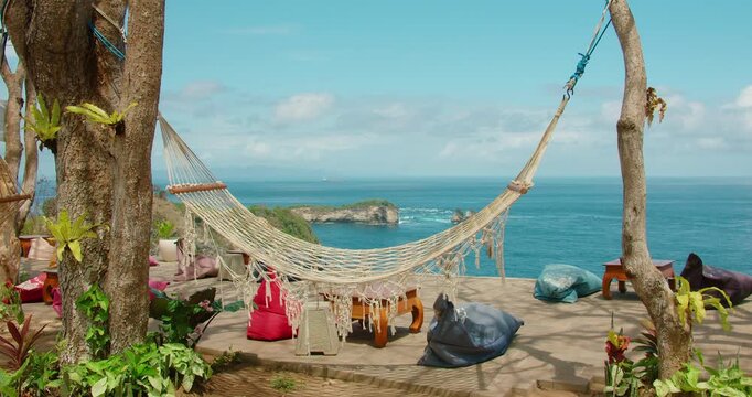 Hammock hangs between trees on clifftop terrace overlooking blue sea in Nusa Penida Bali