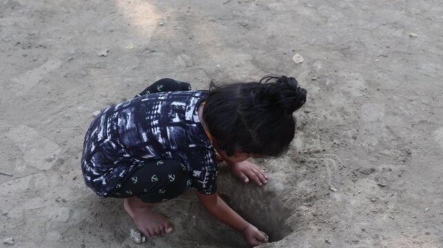 Indian child playing barefoot on the ground, crouching and drawing or digging in the soil with hands, depicting rural childhood, outdoor play, curiosity, innocence, and traditional village lifestyle.