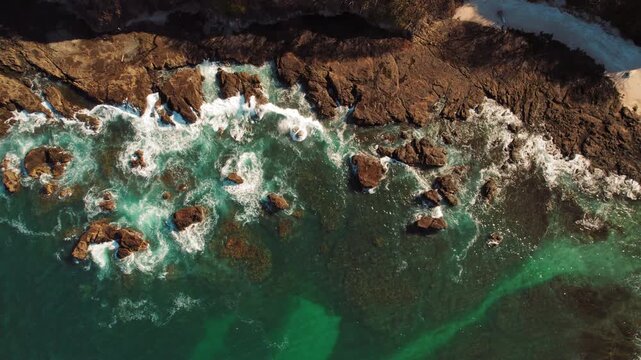 Aerial top down view of turquoise ocean waves hitting dark coastal rocks