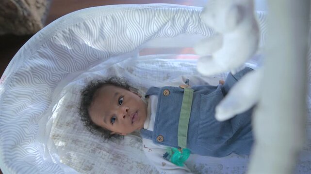 Adorable African American infant boy rests in white mesh bassinet with pacifier clipped to blue outfit while a soft plush mobile toy turns above him and the curious kid watches quietly.