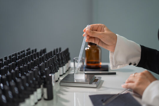 Female perfumer is working on the fragrance: a measuring cup is on the scales and a hand with a pipette is pouring liquid, there are many bottles and bottles around