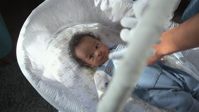 African American infant rests in soft bassinet at home while parent hand adjusts blue outfit and white plush toy dangles above, adorable babe looking up with wide curious eyes in warm daylight.
