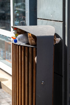 Overfilled public trash can on a city street with plastic bottles and paper waste. Environmental pollution concept. Modern wooden and metal dustbin full of garbage outdoors. Urban waste management 