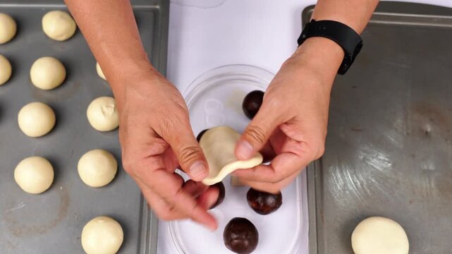 Chef's hands shaping white dough around dark chocolate filling to prepare homemade round cookies or buns on a white surface, with other dough balls on baking trays