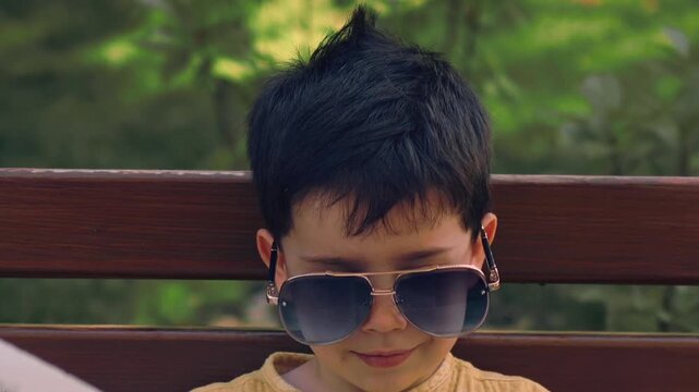 Cute little boy wearing sunglasses and a yellow shirt walking in a sunny green park during summer. Happy childhood and outdoor lifestyle portrait of a fashionable child enjoying nature.