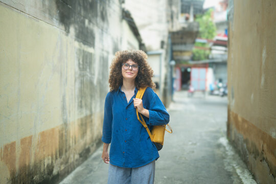 Portrait of Caucasian Woman Traveler with Curly Hair and Yellow Backpack in Asian Alley Street