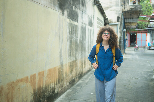 Caucasian woman traveler with backpack walking through an old Asian alleyway smiling with curly hair and glasses