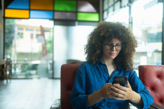 Caucasian Woman with Curly Hair and Glasses Using Smartphone in Colorful Cafe