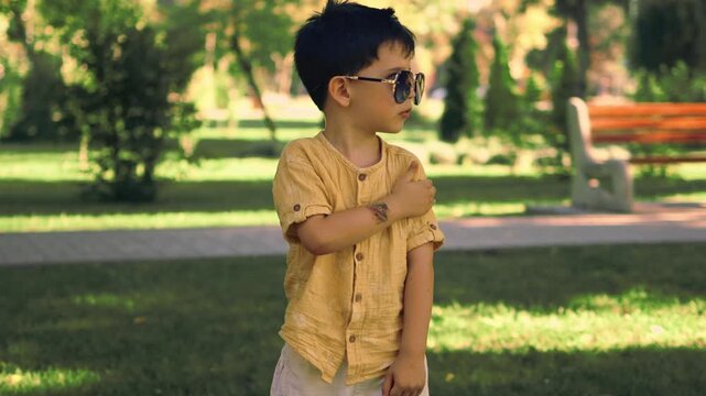 Cute little boy wearing sunglasses and a yellow shirt walking in a sunny green park during summer. Happy childhood and outdoor lifestyle portrait of a fashionable child enjoying nature.