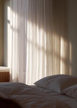 Bedroom with a bed in the foreground. the bed has white sheets and two white pillows. the window is covered with sheer white curtains that are open, creating a soft and airy atmosphere.
