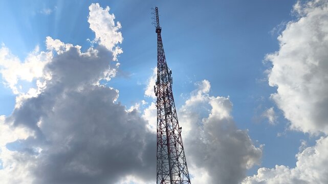 Cellular communication tower rising into a clear blue sky with puffy clouds, featuring multiple antennas and modern infrastructure for wireless networks, mobile connectivity, telecommunications.