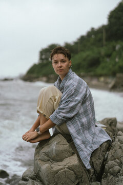 Woman with pixie cut sits on rocks by the sea