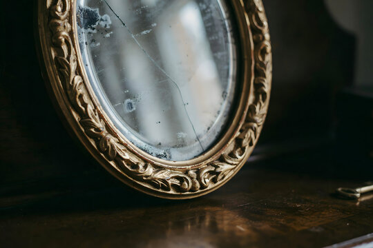 An antique, oval-shaped mirror with an ornate, carved gold frame, showing signs of age, cracks, and oxidation, resting on a dark wooden surface