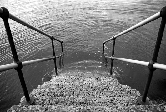 Concrete steps with a metal railing descend into rippling water at high tide in Harwich, Essex, captured in black and white opposite the container terminal, creating a symmetrical composition