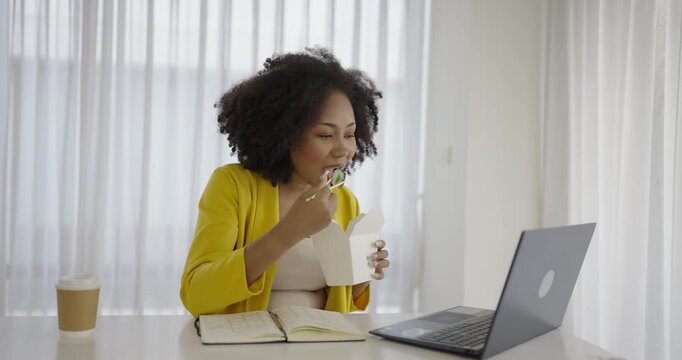 Portrait of young African woman enjoying Chinese food in office lunch break, female employee holding chopsticks eating takeaway Chinese or Korean noodles lunch box meal at workplace in home office.
