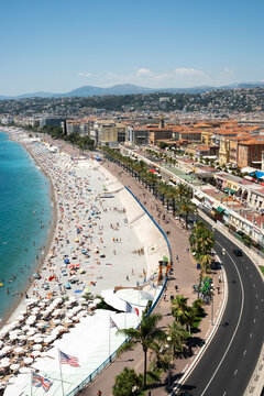 A vibrant view of the Promenade des Anglais and Nice's pebble beach, featuring palm trees, flags, and the turquoise Mediterranean Sea