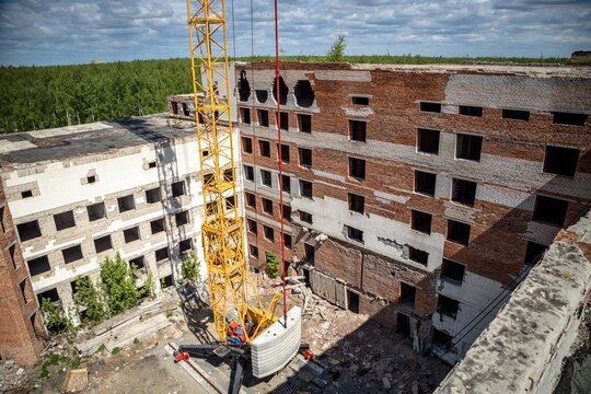 Aerial view of construction crane and abandoned brick building with broken windows, demolition site and forest background, urban decay and industrial scene