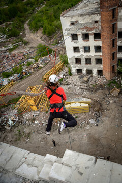 Woman in red jacket and helmet rope jumping with legs kicked back near abandoned brick building, extreme sport adrenaline and thrill concept