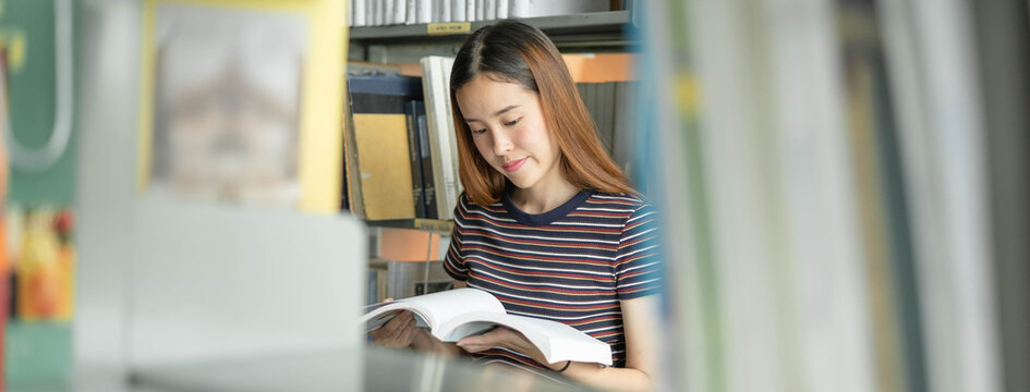 Young beautiful Asian female student reading a textbook and doing research in the university library background with selective focus on bookshelf blur.