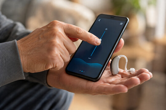 Senior man adjusting a hearing aid with a smartphone app, illustrating digital hearing care, assistive technology, accessibility, connected healthcare and modern hearing loss management