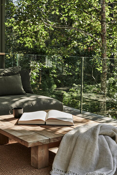Open hardback book on a chunky teak table beside a folded blanket and coffee cup on a terrace in a serene forest retreat setting