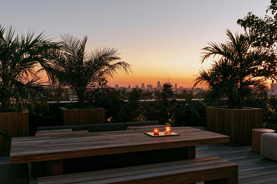 Urban rooftop garden with wooden dining table and candles, surrounded by lush greenery at sunset with city skyline in the background