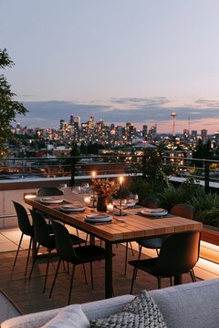 Teak dining table set on urban rooftop with matte black chairs, city skyline illuminated at dusk, warm ambient lighting enhancing the scene
