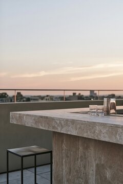 Monolithic travertine bar counter on urban rooftop with brushed black metal stool and glassware reflecting warm dusk light