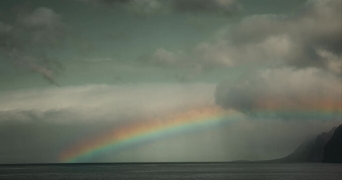 Ocean landscape with volcanic rocks and bright rainbow on cloudy sky. Los Gigantes cliffs in Tenerife.