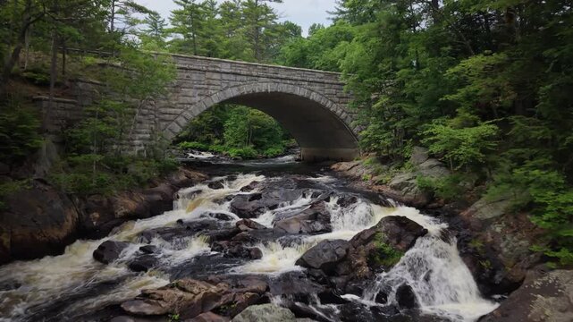 Dramatic drone fly over Profile Falls rushes beneath the stone Route 3A arch bridge surrounded by dense green forest in a rocky gorge, Bristol, New Hampshire, USA