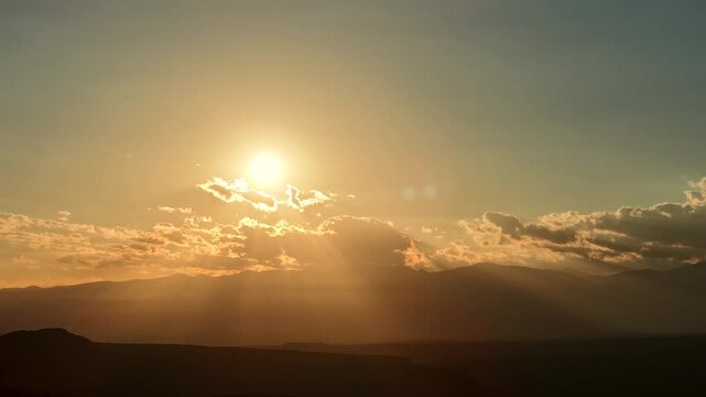 4K drone timelapse of the golden morning sun breaking above a band of scattered clouds. Radiant crepuscular rays ripple downward, illuminating the hazy, silhouetted Sangre de Cristo mountain range.