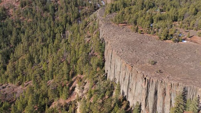 4K aerial view of a sheer volcanic cliff face featuring striking columnar basalt jointing. The massive, vertical grey igneous rock formations stand prominent against a rugged Southwest pine forest.