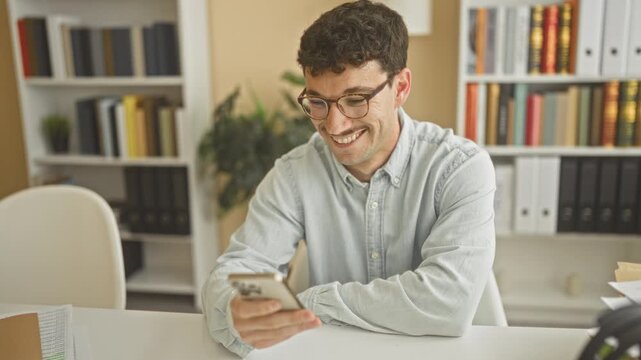 Man holding smartphone and smiling at phone while seated at desk in a building office setting wearing glasses and shirt; work contentment.