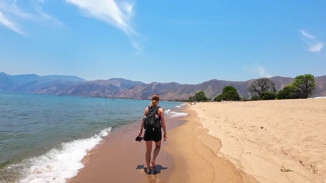 Camera follows a woman walking along golden Usisya Beach beside Lake Malawi, with gentle waves, clear blue sky and scenic mountains on the horizon.