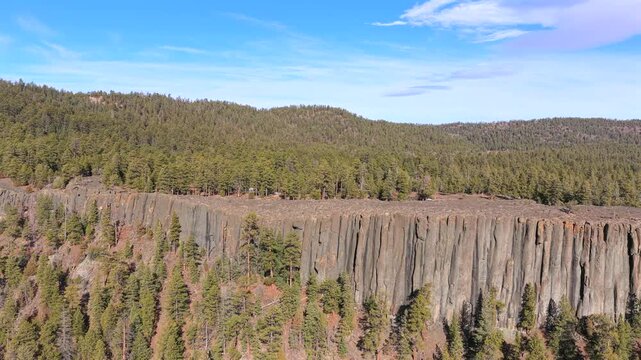 4K aerial scenery showcasing the complex geology of a sheer igneous rock face. Towering grey basalt columns form a dramatic geological boundary surrounded by a Southwest pine forest environment.