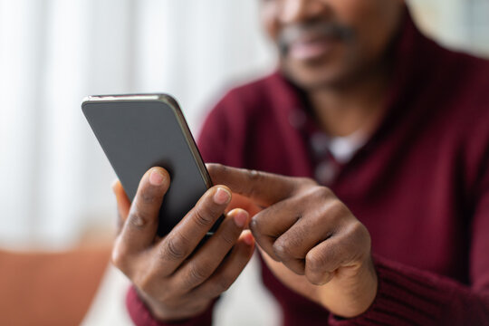 Closeup of mature Black man using smartphone at home, tapping the touchscreen with his finger while browsing, messaging or managing digital tasks in a modern interior