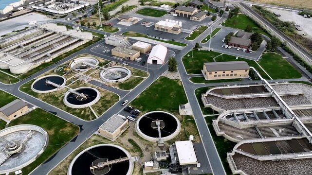 Aerial footage showcasing an extensive wastewater treatment facility with round water tanks, settling filtration vats, skimmers, green lawns, and interconnecting paved roads under natural daylight.