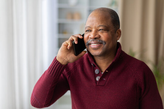 Smiling mature Black man talking on a smartphone at home, enjoying a friendly phone conversation while standing in a bright, comfortable modern living room interior