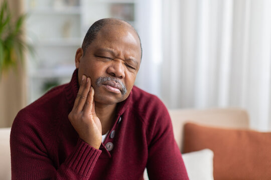 Mature black man holding cheek in pain from toothache, wearing maroon sweater at home, illustrating dental discomfort, oral health and need for care