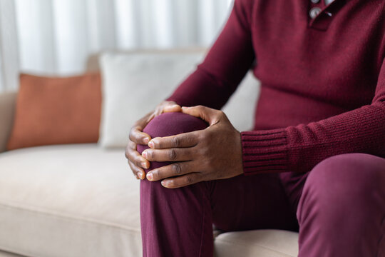Close-up of mature black man holding knee in discomfort while sitting on sofa at home, illustrating joint pain, arthritis, injury, mobility problems and physical discomfort