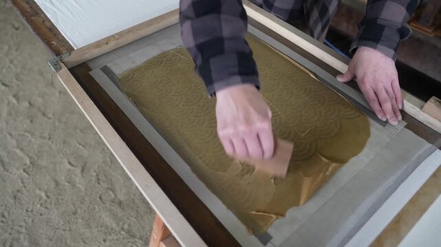 Wooden spatula drags khaki resist paste across the wave stencil, pressing it through the mesh onto the cloth underneath