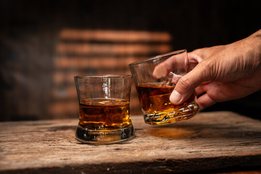 Hand Holding Glass of Whiskey with Ice on Wooden Table in Bar