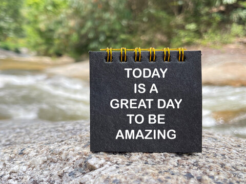 Inspirational message reading &ldquo;Today Is a Great Day to Be Amazing&rdquo; displayed on a small black notepad beside a flowing stream in nature.